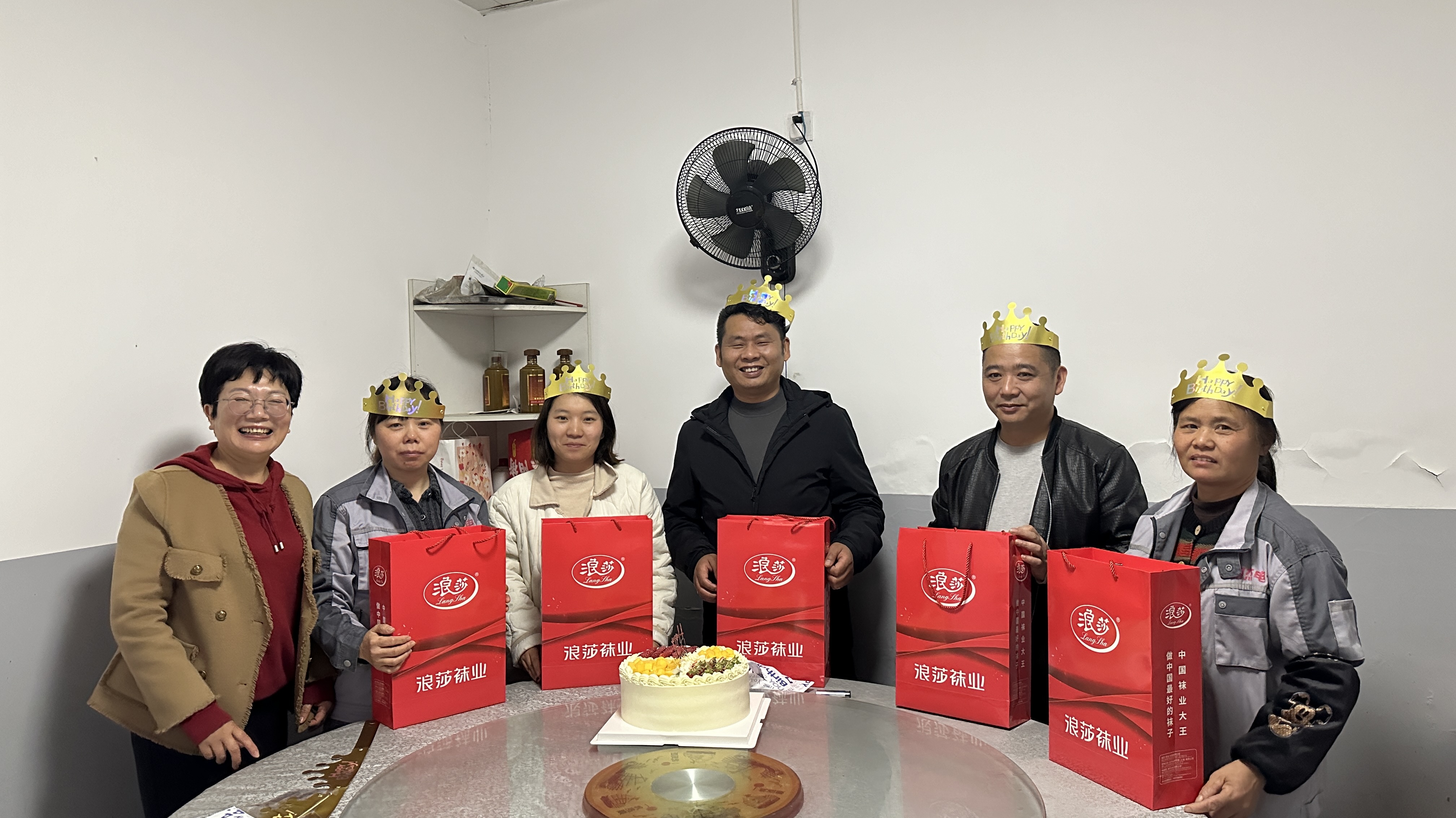Employees gathered around a birthday cake, some wearing birthday hats and holding gift bags